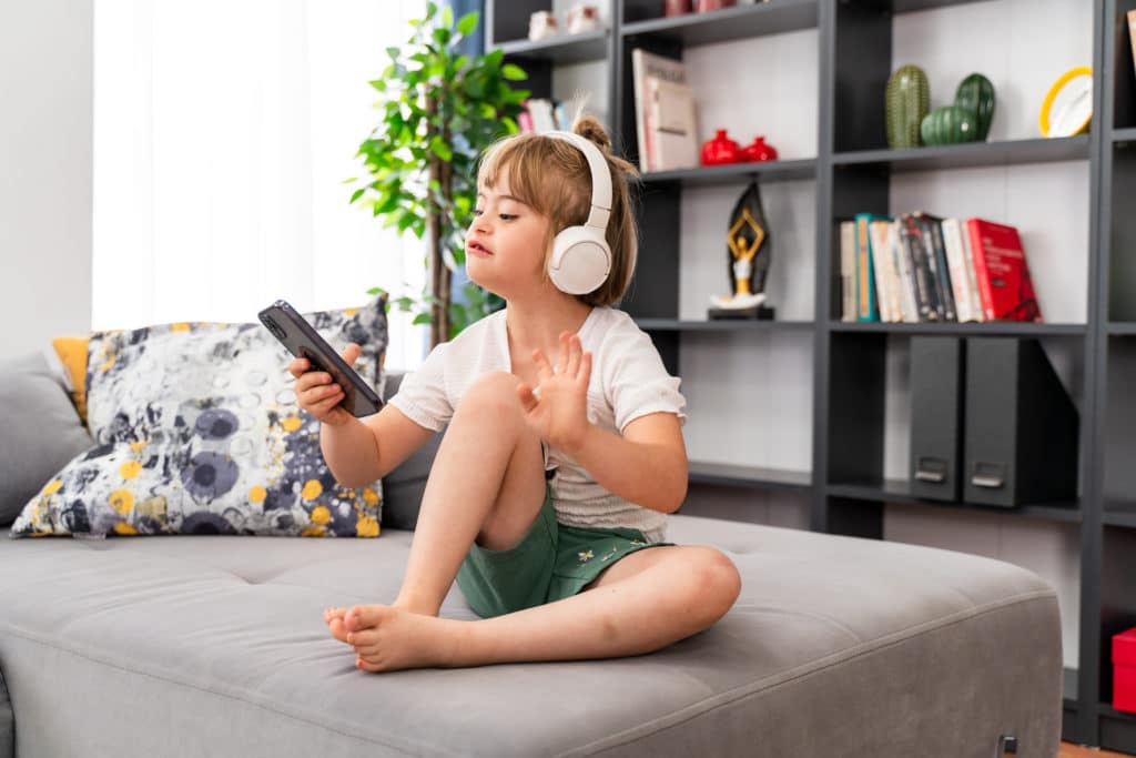 Portrait of girl with Down syndrome using smartphone. Diversity kid listening to music in headphones sitting on sofa at home. Audio development for children with disabilities, inclusive education